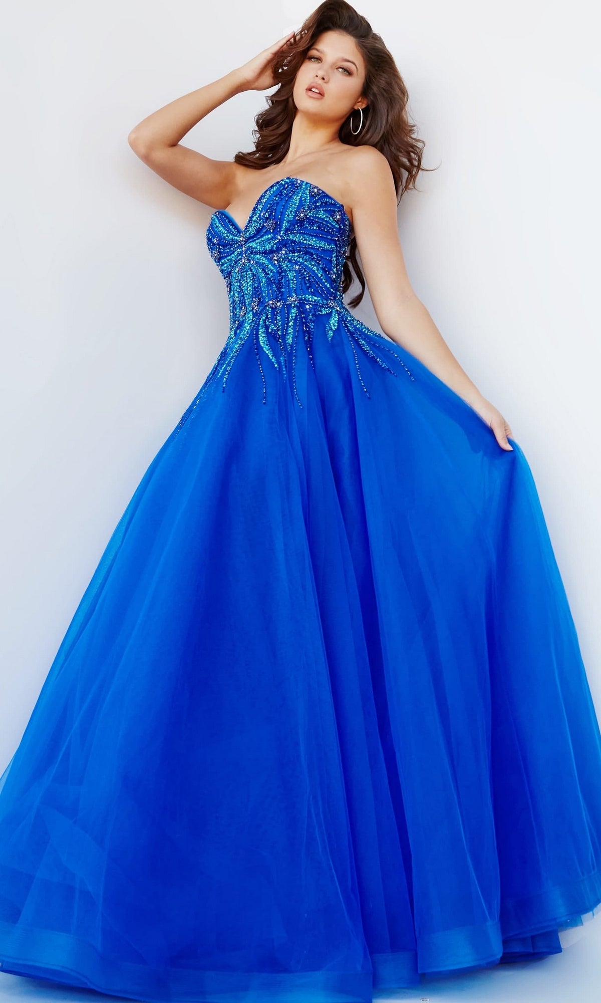 A woman wearing the Jovani Strapless Sweetheart Long Prom Gown 07946, adorned with beaded embellishments, stands against a light background, holding out the skirt with one hand.
